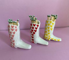 three white boots with hearts and pencils in them on a pink surface, one is made out of ceramic