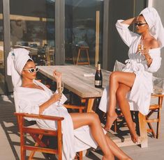 two women in white robes sitting at a wooden table with wine glasses on their feet