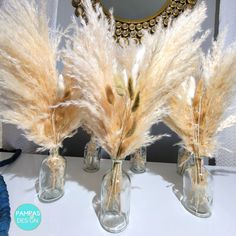 three glass vases with dried grass in them on a table next to a mirror