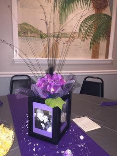 a table with purple flowers in a black box on it and a palm tree behind it