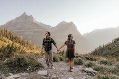 a man and woman holding hands while walking on a trail in the mountains with trees