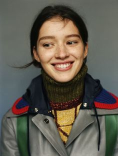 a woman with a jacket and scarf on smiling at the camera while standing in front of a gray wall