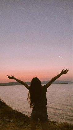 a woman standing on top of a hill next to the ocean with her arms outstretched