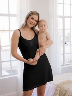 a woman holding a baby in her arms and smiling at the camera while standing next to a bed