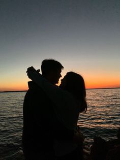 a man and woman standing next to each other near the ocean at sunset with their arms around each other