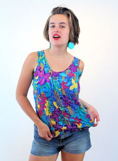 This is a colorful 90s vintage "Gianna" tropical print tank top! Made of soft rayon, it's printed with bold royal purple, magenta and yellow lilies against an ocean blue and purple background. This top has 1.5" wide shoulder straps and a scoop neck in the front and back. It pulls over your head with no zippers or buttons and the hem comes to your waist. This shirt is a vibrant warm weather piece, perfect for summer. This garment is in very good, freshly laundered, clean condition with no flaws or wear noted. The label reads "Gianna, M, Made in Mexico, 100% Rayon, Machine wash cold, Tumble dry low." While it is labeled as a Medium, it fits like a Women's Small according to the measurements: Chest - 34 inches (36 maximum pulled taut), Waist - 36 inches, Shoulders - 11 inches, Length - 26 inc Colorful Bold Print Top For Summer, Colorful Bold Print Summer Top, Colorful Top With Bold Print For Summer, Tropical Sleeveless Tops With Floral Print, Tropical Sleeveless Floral Print Top, Retro Multicolor Tank Top, Casual Purple Tank Top With Floral Print, 90s Floral Print Summer Tops, 90s Style Floral Print Summer Tops