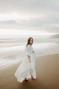 a woman is walking on the beach with her dress blowing in the wind and she's looking at the camera