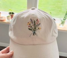 a white baseball cap with flowers embroidered on the front and side, sitting in front of a window