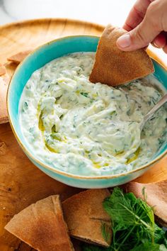 a person dipping dip into a blue bowl with pita chips and parsley on the side