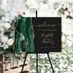 a black and gold wedding welcome sign with flowers in the background