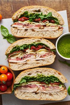 three sandwiches with meat, cheese and veggies next to a bowl of pesto