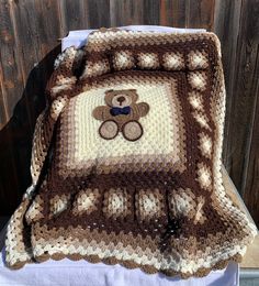 a crocheted teddy bear blanket on top of a white table with a wooden fence in the background