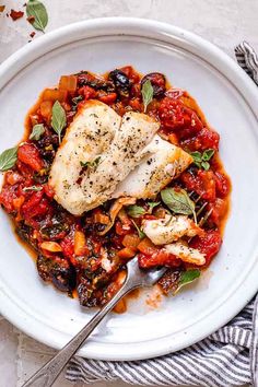 a white plate topped with fish covered in tomato sauce and garnished with herbs