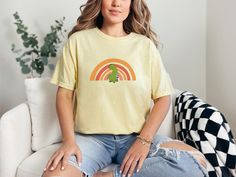 "Cute Dinosaur standing in front of a boho rainbow. Simple and sweet comfy tee. For the oversized look - size up - see size chart for guidance. The photos above show relaxed fit as well as more true to size. Whether you're heading out on a road-trip or hanging out with friends, you'll love the comfortable, oversized fit of this Comfort Colors shirt. The mineral wash feel is super soft and perfect for any casual occasion. Super cute with your favorite jeans, leggings, or just about anything -  you can't go wrong with this super flexible fit -  This is a standard unisex size Comfort Colors Tee.  For an oversized tee, please size up. If you are looking for an oversized \"T-shirt Dress\" look, we recommend sizing up 2 sizes. Please review the size chart to ensure you receive the fit you want." Green Dinosaur Print Tops For Spring, Green Dinosaur Print Top For Spring, Dinosaur Print Short Sleeve Tops For Spring, Cute Dinosaur Print Tops For Spring, Short Sleeve Dinosaur Print Tops For Spring, Short Sleeve Tops With Dinosaur Print For Spring, Spring Short Sleeve Tops With Dinosaur Print, Cute Dinosaur Print Top For Spring, Cute Spring Dinosaur Print Top