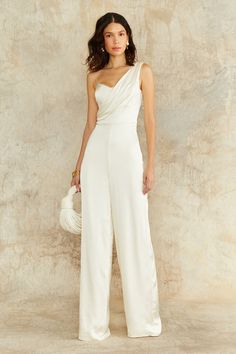 a woman in a white one shoulder jumpsuit holding a white purse and posing for the camera