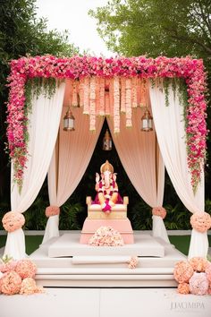 The image showcases a stunning Indian wedding mandap adorned with a vibrant cascade of pink and peach flowers. The mandap, a rectangular structure, is framed by draped white fabric and adorned with hanging lanterns. A traditional Hindu deity, Lord Ganesha, is placed on a pedestal at the center of the mandap. The surrounding area features lush greenery, creating a serene and romantic atmosphere. The overall color theme is soft and feminine, with the pink and peach flowers contrasting beautifully against the neutral backdrop. Peach Indian Wedding Decor, Wedding Stage Decorations Indian Traditional, Hindu Wedding Decorations Indian Theme, Indian Wedding Decor, Neutral Backdrop