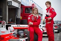 two men in red racing suits sitting next to each other on a race track with cars behind them