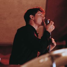 a man holding a microphone up to his mouth while standing next to a drum set