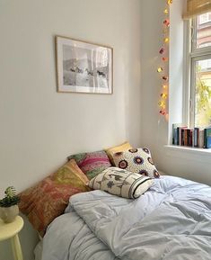 a bed with white sheets and pillows in a small room next to a window filled with books