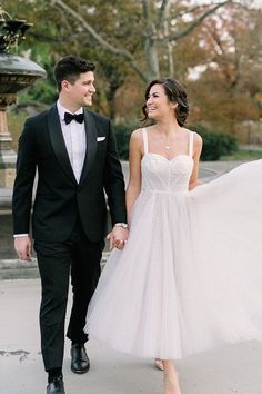 a bride and groom are walking together in the park, dressed in black tuxedos