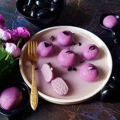 a white plate topped with purple desserts next to pink flowers and black bowls filled with chocolate