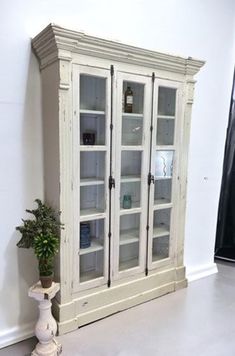 an old white china cabinet with glass doors next to a potted plant on the floor