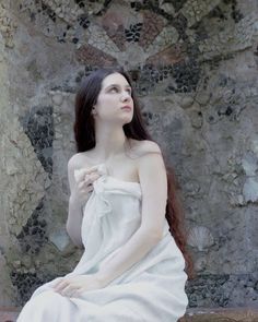 a woman with long hair in a white dress sitting on a stone bench next to a rock wall