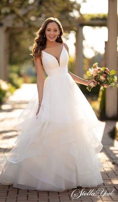 a woman in a white wedding dress holding flowers and smiling at the camera while standing on a brick walkway