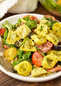 a white bowl filled with pasta and vegetables on top of a wooden table next to a fork