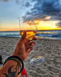 a person holding up a glass of wine on the beach at sunset or sunrise time