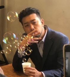 a man sitting at a table blowing soap bubbles on his finger and drinking from a cup