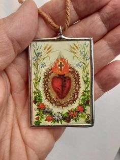 a hand holding a small square pendant with a heart in the center and flowers around it