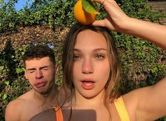 a woman holding an orange on top of her head next to a man who is covering his eyes