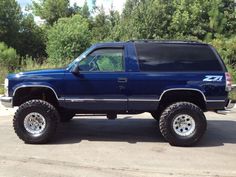 a large blue truck parked on top of a parking lot next to trees and bushes