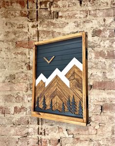 a wooden sign hanging on the side of a brick wall with mountains and trees painted on it