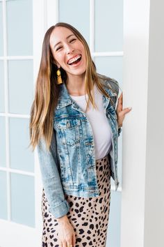 a woman in a leopard print skirt and jean jacket smiles as she leans against a wall