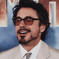 a man wearing glasses and a suit smiling at the camera while standing in front of a movie poster