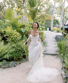 a woman in a white wedding dress walking through the jungle with palm trees behind her