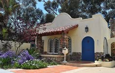 a white house with blue doors surrounded by trees and flowers in front of the building