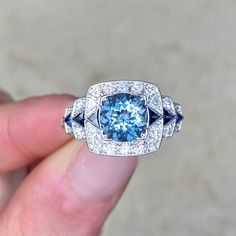 A geometric gemstone ring featuring a 1.22-carat round cut aquamarine set in prongs. Triangular French cut sapphires are set in a row flanking the aquamarine and on the stepped shoulders. Round brilliant cut diamonds are set surrounding the aquamarine and sapphires and continue along the shoulders. The total diamond weight of this ring is approximately 0.45 carats. The sapphires weigh 0.22 carats combined. This ring is handcrafted in platinum, decorated with delicate milgrain and an openwork und Round Cut Aquamarine Gemstone Jewelry, Modern Blue Topaz Jewelry With Center Stone, Modern Blue Topaz Center Stone Jewelry, Light Blue Fine Jewelry With Center Stone, Fine Jewelry Aquamarine Round Cut, Light Blue Diamond Jewelry With Accent Stones, Cushion Cut Blue Topaz Jewelry With Center Stone, Light Blue Center Stone Fine Jewelry, Light Blue Round Cut Diamond Jewelry