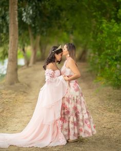 two women in dresses standing next to each other on a dirt road with trees behind them