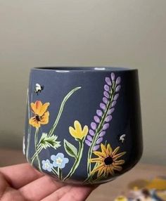 a hand holding up a painted cup with flowers on the outside and bees flying around