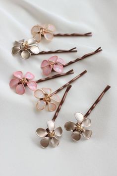 six hair pins with flowers on them sitting on a white cloth covered surface, one is pink and the other is gold