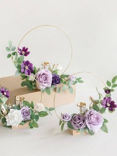 three purple and white flowers are in a box with greenery on it, along with two gold hoop holders