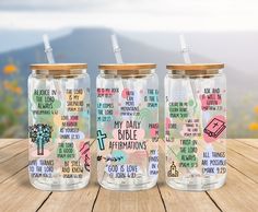 three glass jars with different designs and words on them sitting on a wooden table in front of mountains