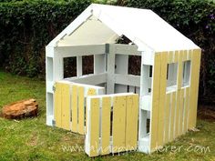 a small yellow and white house made out of wooden pallets in the grass next to a tree stump