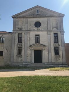 an old white building with a black door and window on the front, surrounded by green grass