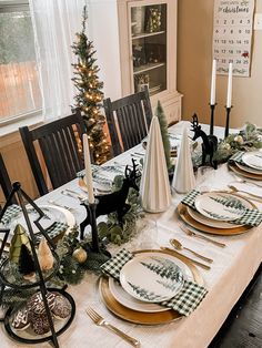 a dining room table set for christmas with plates and place settings, candlesticks and evergreen trees