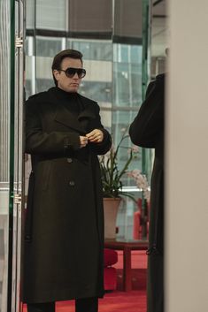 a man in a black coat and sunglasses standing next to a glass wall looking at something