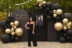 a woman standing in front of a black and gold balloon arch with balloons on it